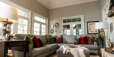 A spacious living room with a gray sectional sofa, colorful cushions, ottoman, and large windows letting in natural light.
